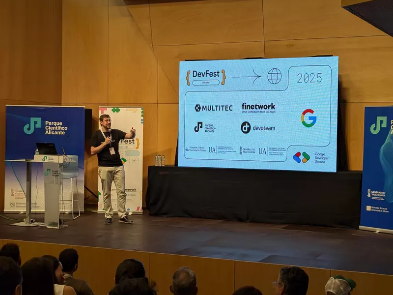 Ponente en el escenario del DevFest Alicante 2025, con el logo de Multitec en el panel de patrocinadores