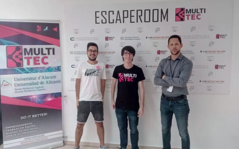 Tres miembros de Multitec ante el panel de la escape room, con el rótulo ESCAPEROOM y el logotipo